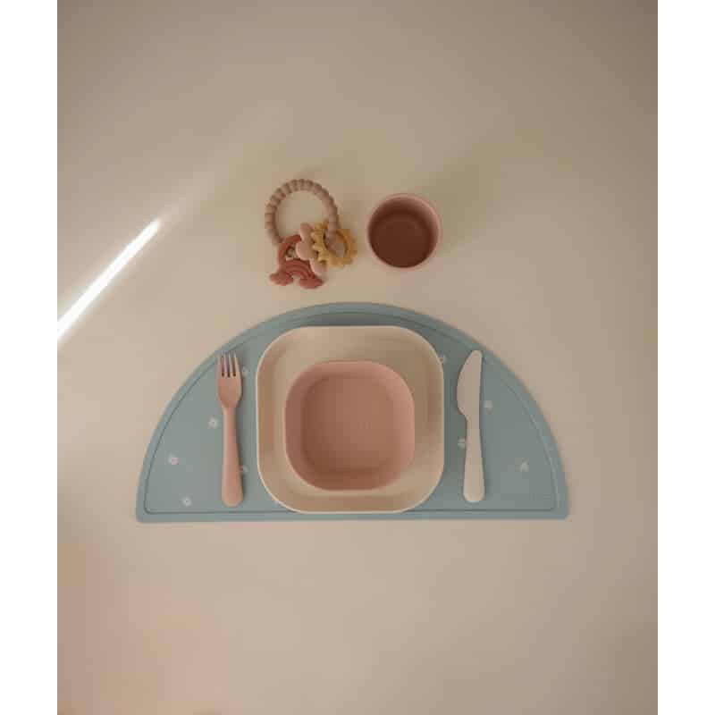 Mushie Dinnerware Cutlery Set in blush and ivory, featuring a bowl, plate, fork, knife, and cup on a blue placemat.