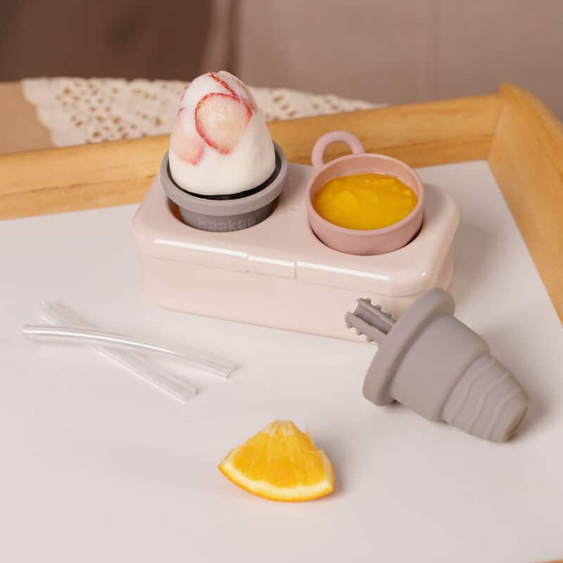 Haakaa Freeze-'n-Sip Ice Cream Mould Set in marigold and slate, featuring a cup, mould, and fruit slice on a tray.