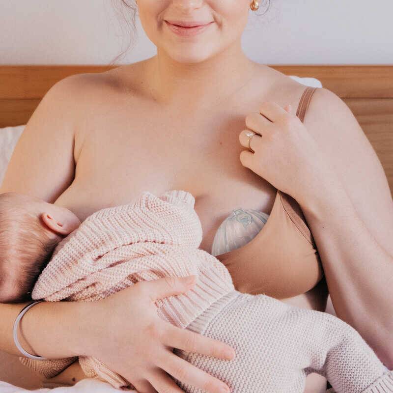 Haakaa Shell Wearable Silicone Breast Pump in use, discreetly worn under a nursing bra while breastfeeding a baby.