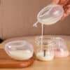 Haakaa Shell Wearable Silicone Breast Pump in clear silicone, pouring milk into a glass collector jar on a wooden surface.