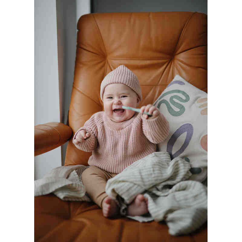 Mushie First Feeding Baby Spoons in blush and Cambridge blue, held by a smiling baby in a cozy chair setting.