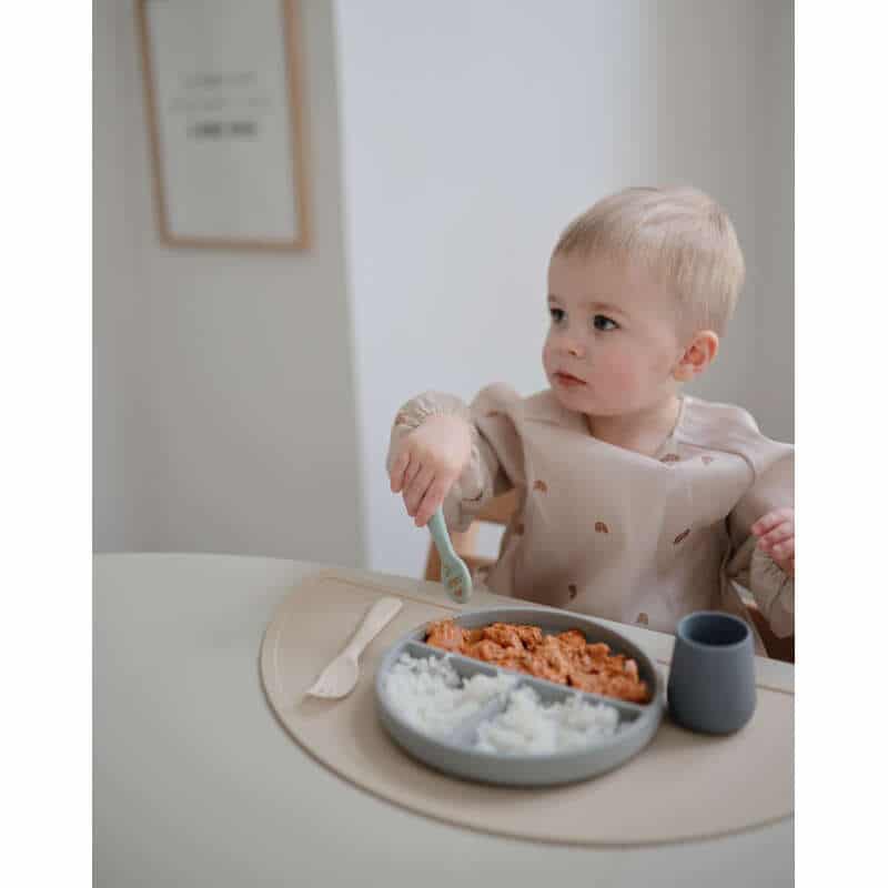 Mushie First Feeding Baby Spoons in blush and Cambridge blue, used by a toddler at a dining table with food.