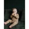 Mushie No-Drop Sun Teether in Sunshine color held by a toddler sitting on a green couch, wearing a beige outfit.
