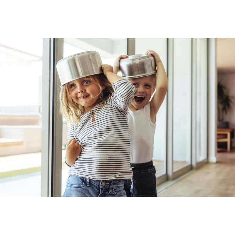 Two children playfully wearing stainless steel pots on their heads, smiling and having fun indoors.