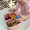 Maccie Munchmate Bento Lunchbox in pink speckles, featuring three compartments with colorful food items, held by a toddler.
