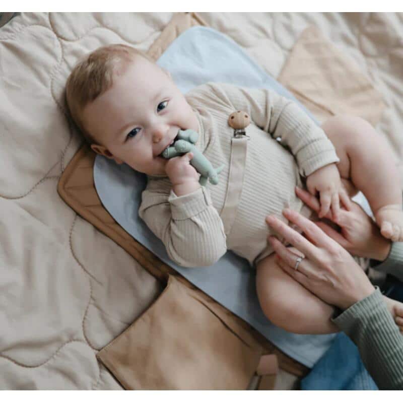 Mushie Figurine Teether in green, held by a baby sitting on a soft quilt, with a parent’s hand gently resting nearby.