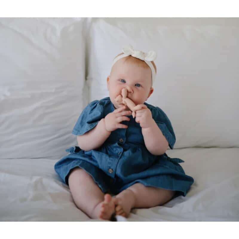 Mushie Figurine Teether in a baby's hands, sitting on a bed, wearing a blue dress and a bow headband.