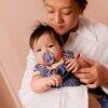 BIBS Studio Colour Pacifier 2 Pack in Baby Blue Dusty Blue Mix and Jasmine Fossil Grey Mix, held by a parent with a baby.