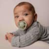 Difrax Dynamic Soother Prime in coconut/pistachio, held by a baby wearing a grey outfit, lying on a soft surface.