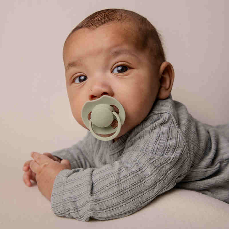 Difrax Dynamic Soother Prime in coconut/pistachio, held by a baby wearing a grey outfit, lying on a soft surface.