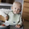 Difrax LOVI Silicone Bib in pistachio worn by a toddler sitting at a table with a bowl of strawberries.
