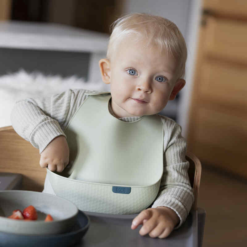 Difrax LOVI Silicone Bib in pistachio worn by a toddler sitting at a table with a bowl of strawberries.