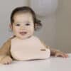 Difrax LOVI Silicone Bib in nude, worn by a smiling baby at a table, showcasing its practical design and soft material.