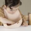 Difrax LOVI Silicone Bib in nude, featuring a soft design with a child exploring it at a table setting.