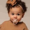 Difrax Dynamic Soother Prime in nude/blush, held by a baby with curly hair, wearing a cozy brown sweater.