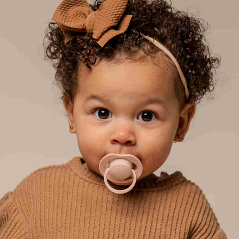 Difrax Dynamic Soother Prime in nude/blush, held by a baby with curly hair, wearing a cozy brown sweater.