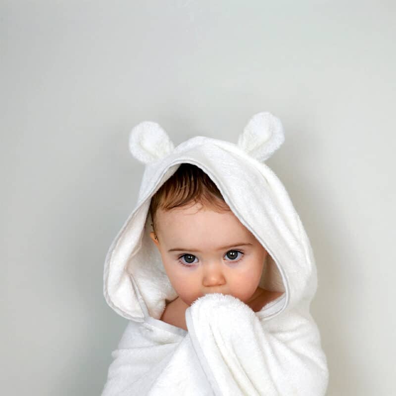 Shnuggle white bamboo wearable baby towel with bear ears, draped over a baby holding the edge, against a light background.