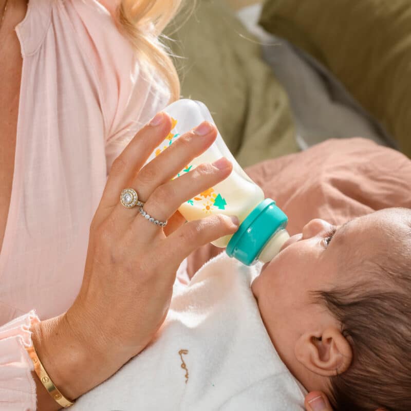 Pigeon Flexible Curved Bottle 250ml in beach color, held by a caregiver feeding a baby, soft bedding in background.