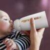 Momcozy Disposable Baby Bottle Kit in white, held by a caregiver while a baby drinks from it, featuring a clear measurement