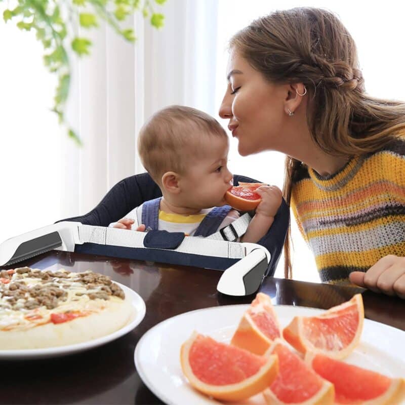 Momcozy Mtwml Portable High Chair in use at a dining table, with a mother and baby enjoying a meal together.