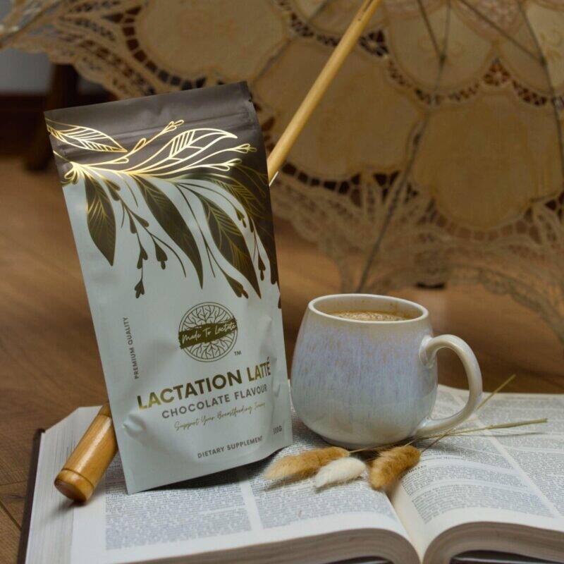 Made to Lactate Enhanced Lactation Chocolate Latté in a decorative pouch beside a cozy mug on an open book.
