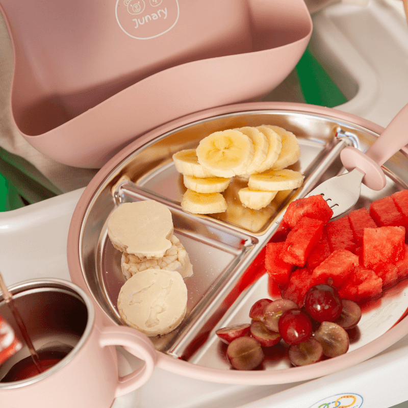 Junary Little Gripper Starter Kit in pink with a divided plate holding banana slices, watermelon, grapes, and cookies.