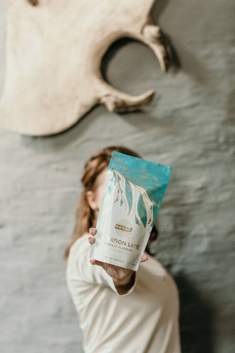 Made to Lactate Lactation Vanilla Latté in a stylish bag held by a woman, set against a rustic wall backdrop.