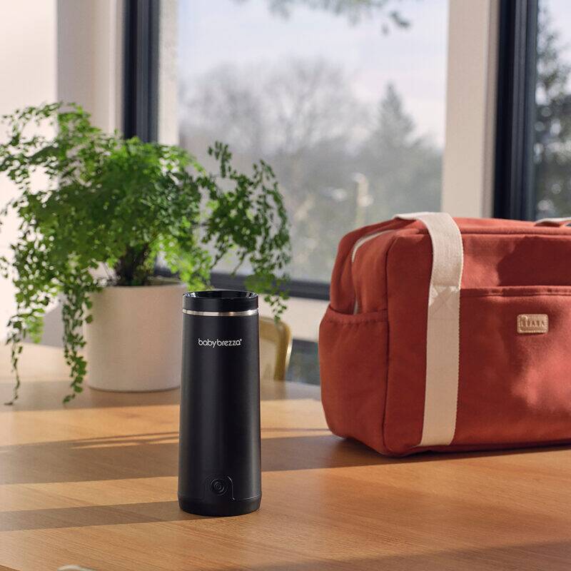 Baby Brezza SuperFast Portable Warmer in sleek black, placed on a wooden table next to a stylish diaper bag and a potted