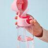 b.box 600ml Tritan drink bottle in pink, featuring a flip-top lid and straw, held in a hand against a light blue background.