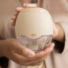 Difrax Handsfree Breastpump in beige held by a person, showcasing its ergonomic design and transparent collection container.