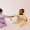 Difrax Crown Teething Ring held by two babies in colorful outfits, sitting on a light background, engaging playfully.
