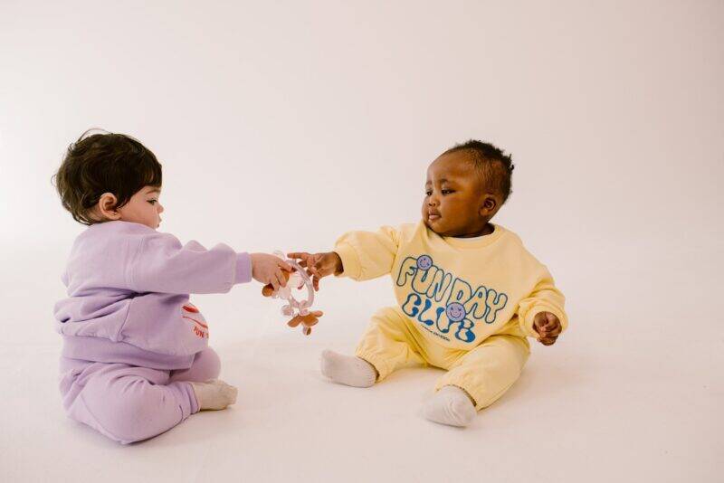 Difrax Crown Teething Ring held by two babies in colorful outfits, sitting on a light background, engaging playfully.