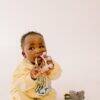 Difrax Crown Teething Ring held by a baby in a yellow outfit, sitting on a light background with a toy nearby.