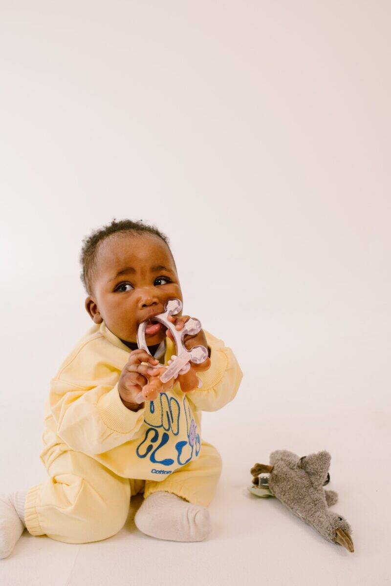 Difrax Crown Teething Ring held by a baby in a yellow outfit, sitting on a light background with a toy nearby.