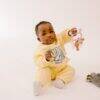 Difrax Crown Teething Ring held by a baby in a yellow outfit, sitting on a light background with a toy nearby.