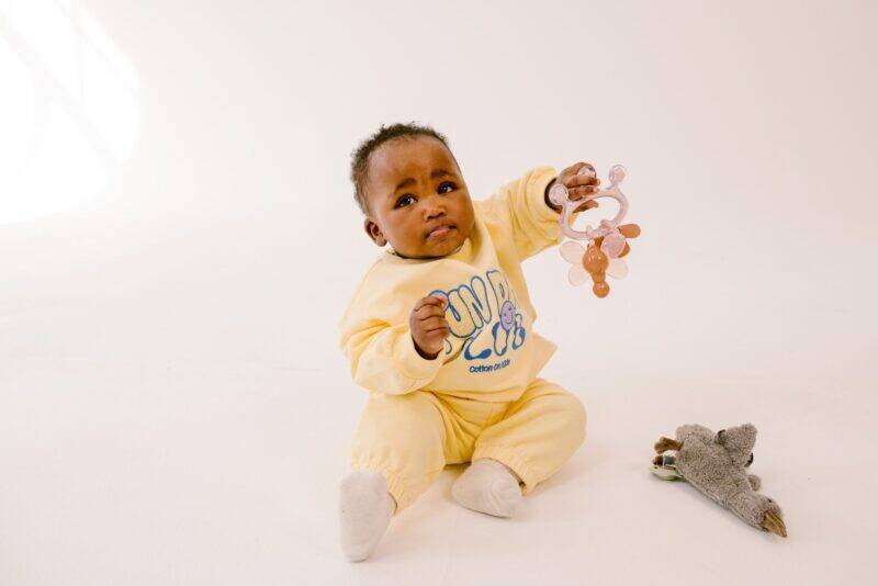 Difrax Crown Teething Ring held by a baby in a yellow outfit, sitting on a light background with a toy nearby.