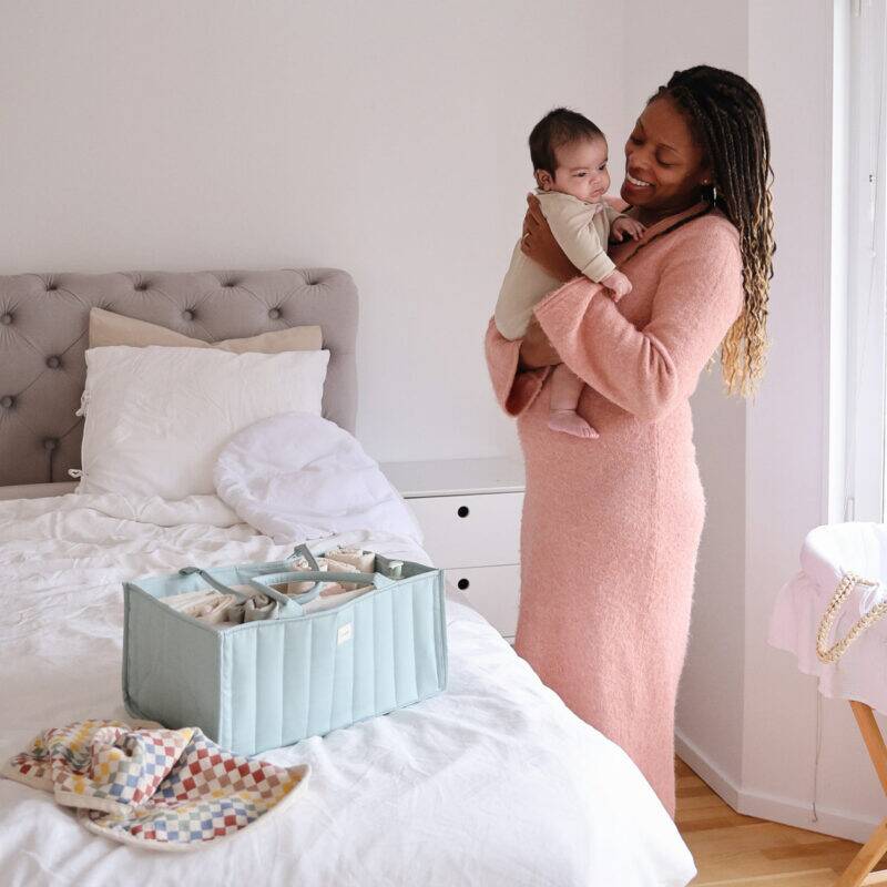 Mushie Diaper Caddy in Blue Flowers and Blush, placed on a bed, with a mother holding her baby nearby.