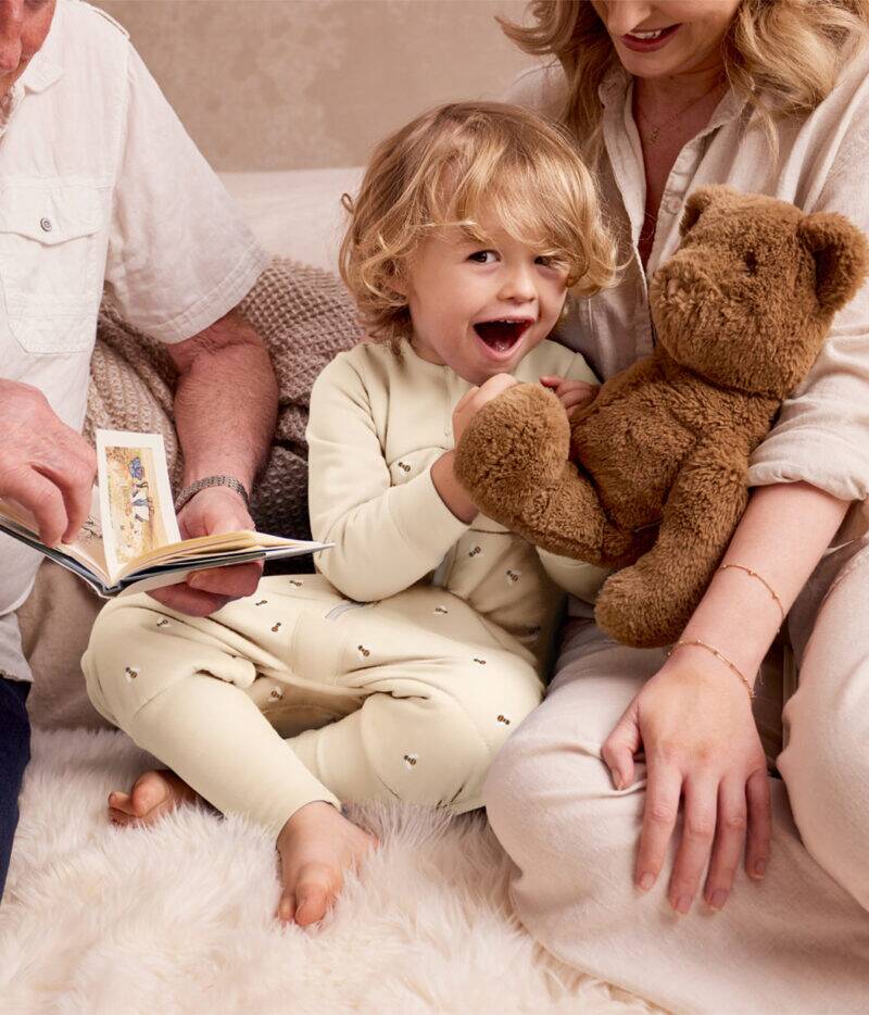 Love to Dream Sleep Suit Long Sleeve 2.5 TOG in Oatmeal Bees, child smiling while holding a teddy bear with family.