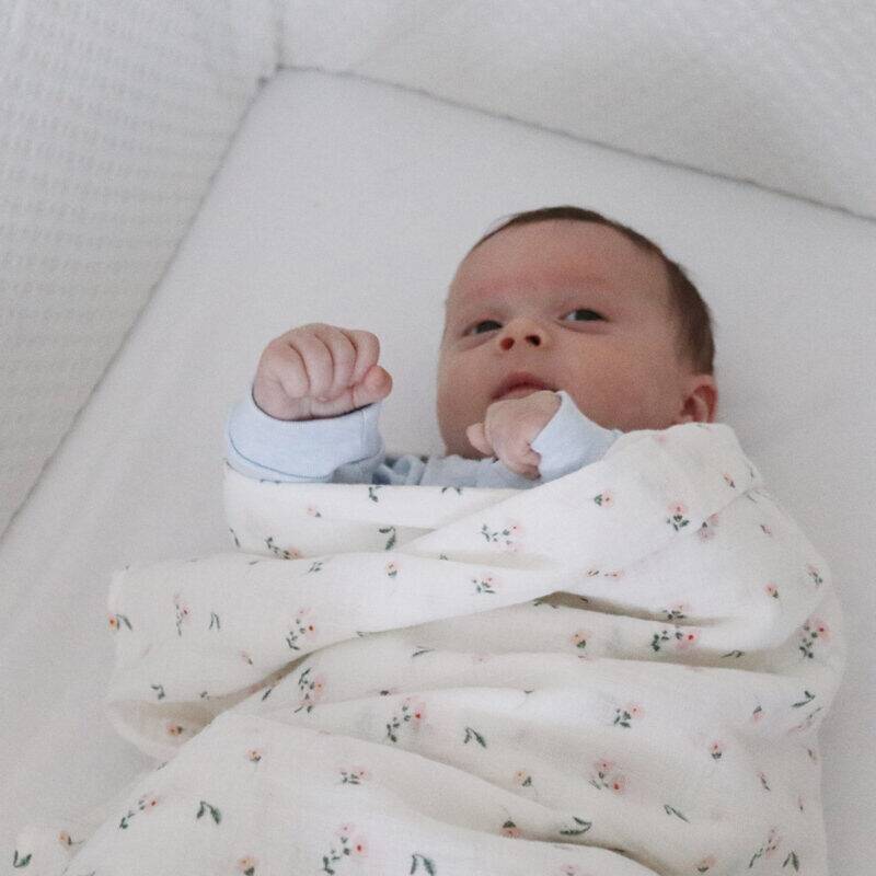 Snuggletime Deluxe Muslin Receiver in pink blossoms, wrapped around a baby lying on a white crib mattress.