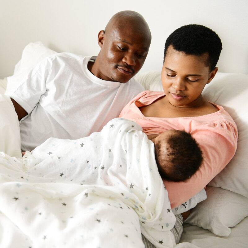 Snuggletime Bamboo Muslin Receiver in blue star, wrapped around a baby, with parents lovingly watching in a cozy bedroom