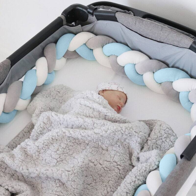 Snuggletime Braided Cot Bumper in blue, grey, and pink, surrounding a sleeping baby wrapped in a cozy blanket.