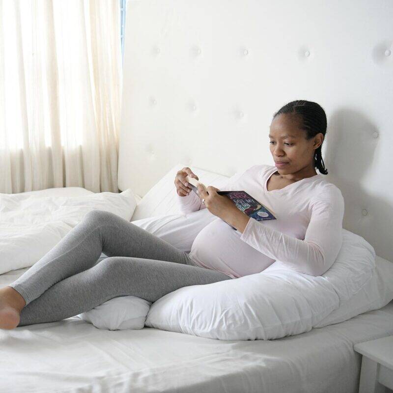 Snuggletime Body Comfort Pillow in grey, taupe, and white, shown in a cozy bedroom setting with a pregnant woman reading.