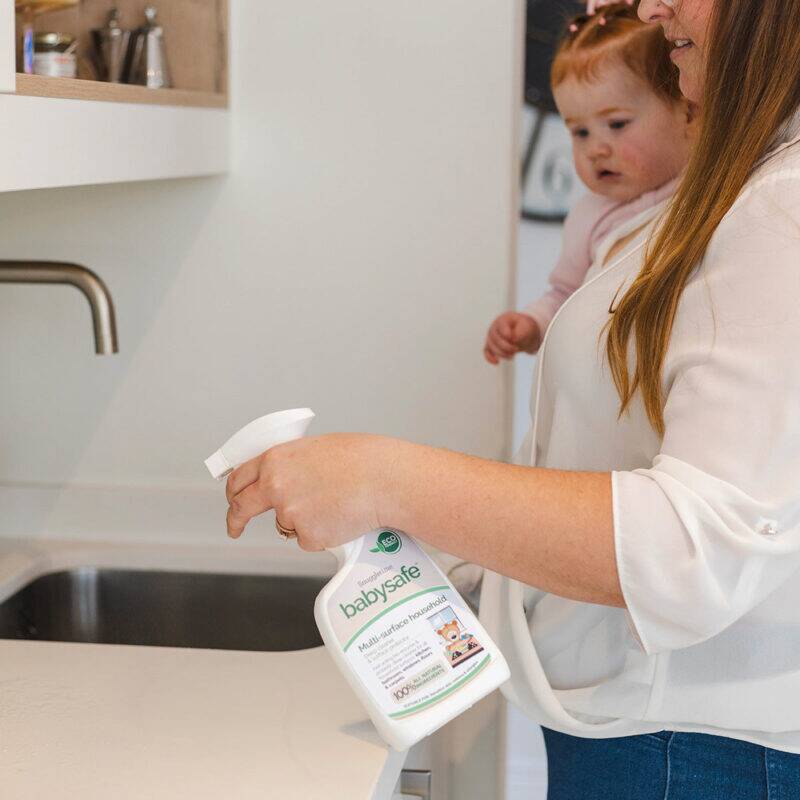 BabySafe Multi-Surface Household Deep Cleaner in a 500ml bottle, used by a woman while holding a baby, cleaning a kitchen