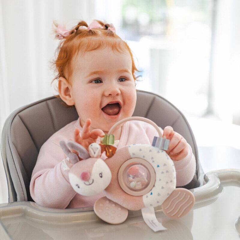 Back To Nature Teether & Spinning Rattle in blue and pink, held by a smiling baby in a high chair.