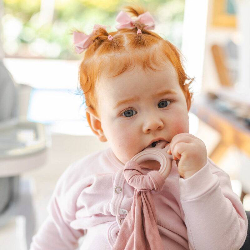 Beige and pink Back To Nature silicone teether muslin blankie held by a baby with red hair, sitting in a bright room.