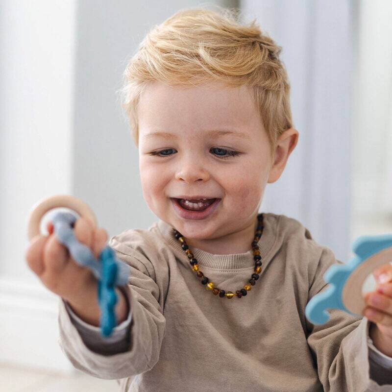 Back To Nature Teether Ring in blue and pink, held by a smiling toddler wearing a necklace, in a bright indoor setting.