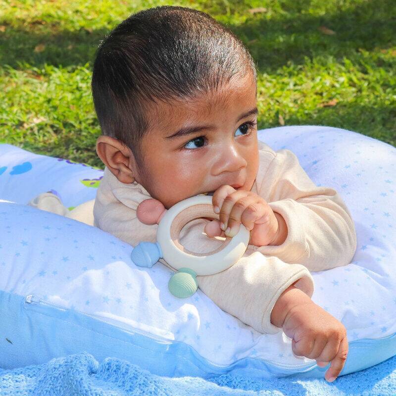 Back To Nature Silicone Chewable Teether with Wooden Ring, featuring soft pastel colors, held by a baby on a grassy surface.