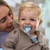 Back To Nature Silicone Pacifier Clip in blue, attached to a smiling toddler, with a mother in a cozy indoor setting.