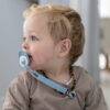 Back To Nature Silicone Pacifier Clip in blue attached to a pacifier, worn by a toddler with curly hair in a cozy indoor