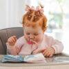 Back To Nature Silicone Pacifier Clip in pink, attached to a baby’s pacifier, as a toddler explores a soft book in a high